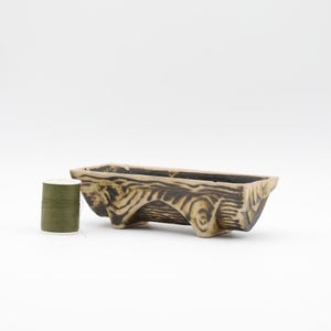 May include: A rectangular, ceramic planter with a wood-grain design in shades of brown and beige. A spool of olive green thread sits to the left of the planter. The planter is approximately 8 inches long.