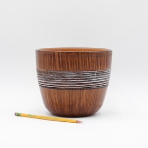 May include: A brown ceramic bowl with a textured wood grain appearance and a band of silver-colored horizontal lines. A yellow pencil with a green eraser lies next to the bowl. The bowl has a rounded shape and is set against a white background.