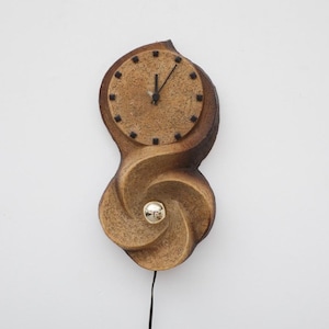 May include: A unique, handcrafted wall clock with a swirling, organic design. The clock face is circular with black markers, and the hands point to 12 o'clock. The clock is made of a light brown material and features a silver-colored pendulum.