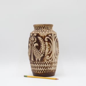 May include: A brown and beige ceramic vase with a textured surface and intricate carved designs. The vase features a repeating pattern of geometric shapes and stylized birds. A yellow pencil rests near the base of the vase.