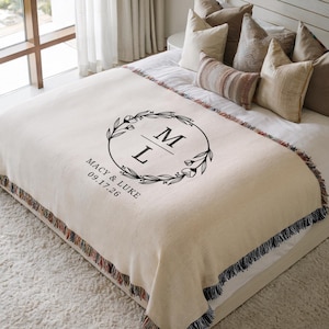 Pode incluir: Cobertor creme com franjas decorativas. O cobertor apresenta um design de coroa circular preta com as letras "M" e "L" no interior. Abaixo da coroa está o texto "Macy & Luke 09.17.26". O cobertor está em uma cama com travesseiros.