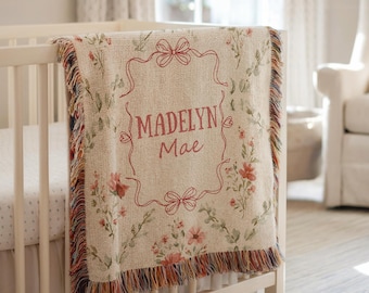 May include: A beige baby blanket with a floral pattern and the name "Madelyn Mae" embroidered in red. The blanket has a fringed edge and is draped over a white cot. The nursery setting includes a white armchair and curtains.