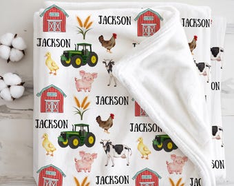 May include: White baby blanket with a farm animal pattern. The blanket features red barns, green tractors, and illustrations of chickens, pigs, cows, and ducks. The name "JACKSON" is printed across the blanket.
