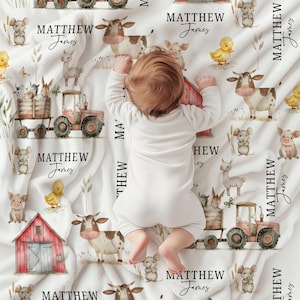 May include: A white baby blanket with a farm animal theme. The blanket features illustrations of cows, pigs, ducks, tractors, and barns. The name "MATTHEW James" is printed repeatedly on the blanket. A baby in a white onesie is lying on the blanket.