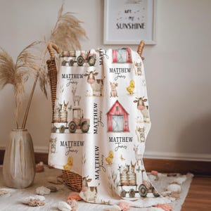 May include: A cream-colored blanket featuring a farm animal theme with illustrations of cows, pigs, and tractors. The blanket has the name "MATTHEW James" printed on it. The blanket is folded in a wicker basket.