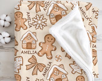 May include: A beige blanket featuring a repeating pattern of gingerbread men, gingerbread houses, snowflakes, and bows. The name "AMELIA" is printed throughout the design. The blanket has a white, plush underside.