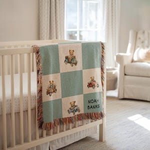 Pode incluir: Um cobertor de bebê com um padrão xadrez com ursinhos de pelúcia dirigindo carros antigos. O cobertor é em tons de verde e bege, com uma borda com franjas. O nome "NOAH BANKS" está impresso.