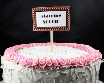 May include: A white cake with pink frosting and a sign that says "starring Sophie".