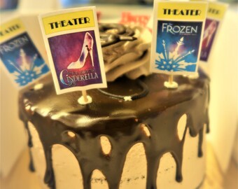 May include: A chocolate cake with a white frosting base decorated with chocolate drizzle and topped with four small flags. The flags are for the Broadway shows Frozen, Cinderella, and two for the generic "Theater" logo.