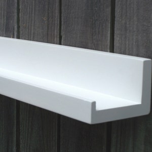 May include: A white floating shelf with a simple, modern design. The shelf is mounted to a wood wall and has a slight lip on the front edge.