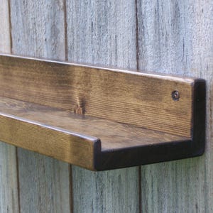 May include: A dark brown wooden shelf with a slightly rounded edge. The shelf is mounted to a light gray wood wall.