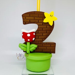 May include: A handmade birthday candle shaped like the number 2, with a brick pattern, a yellow star, and a red and white Piranha Plant. The candle sits on a green pot and includes a yellow candle. The text "Mundo Clay Handmade" is visible.