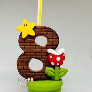 May include: A handmade birthday candle holder featuring the number 8 in a brick design, a yellow star, and a red and white spotted Piranha Plant. A yellow and white striped candle is inserted into the top. The base is a green pipe.