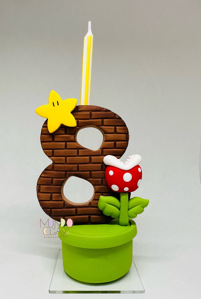 May include: A handmade birthday candle holder featuring the number 8 in a brick design, a yellow star, and a red and white spotted Piranha Plant. A yellow and white striped candle is inserted into the top. The base is a green pipe.