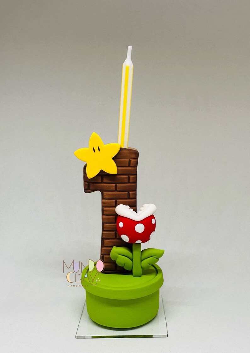 May include: A birthday cake topper with a brown brick-effect number one, a yellow star, and a red and white spotted Piranha Plant. A yellow and white striped candle is inserted into the number one. The topper is on a green pot.