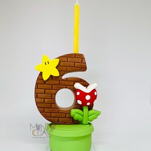 May include: A number six birthday candle with a brick pattern, a yellow star, and a red and white flower. The candle is yellow and sits atop the number. The base is a green pot. The logo "Mundo Clay Handmade" is visible.