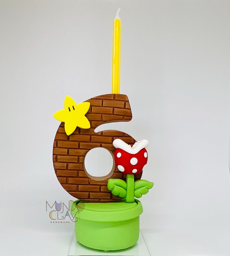 May include: A number six birthday candle with a brick pattern, a yellow star, and a red and white flower. The candle is yellow and sits atop the number. The base is a green pot. The logo "Mundo Clay Handmade" is visible.