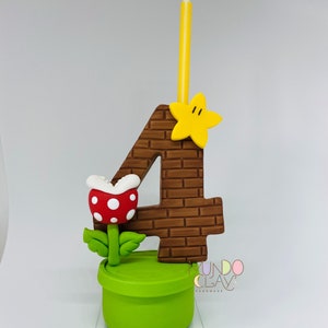 May include: A handmade birthday cake topper. The number 4 is brown with a brick pattern, a yellow star, and a yellow and white striped candle. A red and white spotted Piranha Plant sits in a green pipe. The base is a green cylinder.
