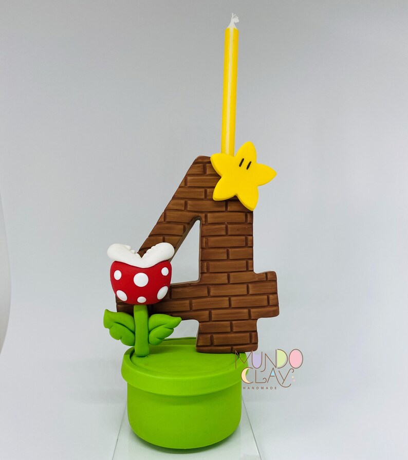 May include: A handmade birthday cake topper. The number 4 is brown with a brick pattern, a yellow star, and a yellow and white striped candle. A red and white spotted Piranha Plant sits in a green pipe. The base is a green cylinder.