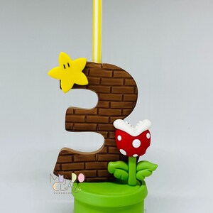 May include: A handmade number three birthday candle with a brick-effect design, a yellow star, and a red and white Piranha Plant. The candle is set in a green pot and has a yellow and white striped candle.