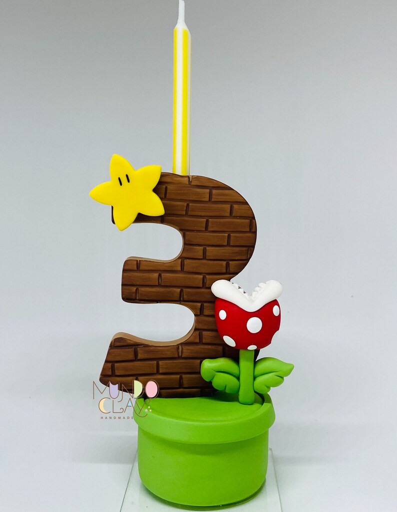 May include: A handmade number three birthday candle with a brick-effect design, a yellow star, and a red and white Piranha Plant. The candle is set in a green pot and has a yellow and white striped candle.