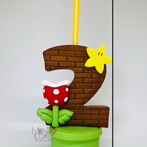 May include: A handmade birthday candle in the shape of the number 2, with a brick design, a yellow star, and a red and white flower. The candle is 15 cm tall and has a yellow candle. The base is a green pot.