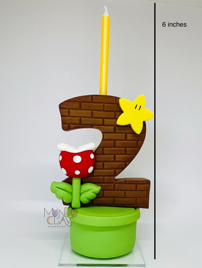 May include: A handmade birthday candle in the shape of the number 2, with a brick design, a yellow star, and a red and white flower. The candle is 15 cm tall and has a yellow candle. The base is a green pot.