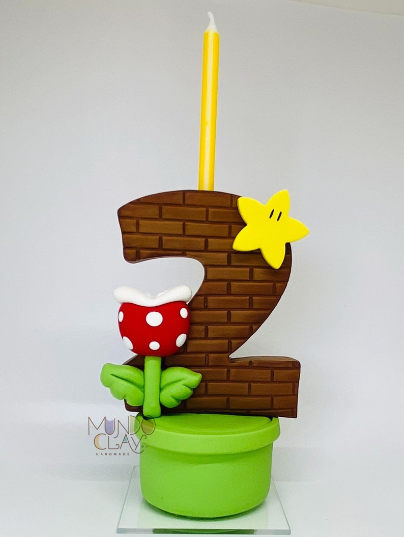 May include: A handmade birthday candle shaped like the number 2, with a brick pattern, a yellow star, and a red and white Piranha Plant. The candle sits on a green pot and includes a yellow candle. The text "Mundo Clay Handmade" is visible.
