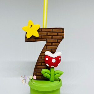 May include: A number seven birthday candle with a brick pattern, a yellow star, and a red and white Piranha Plant. The candle is yellow and white. The base is a green pot. The design is inspired by the Super Mario video game series.