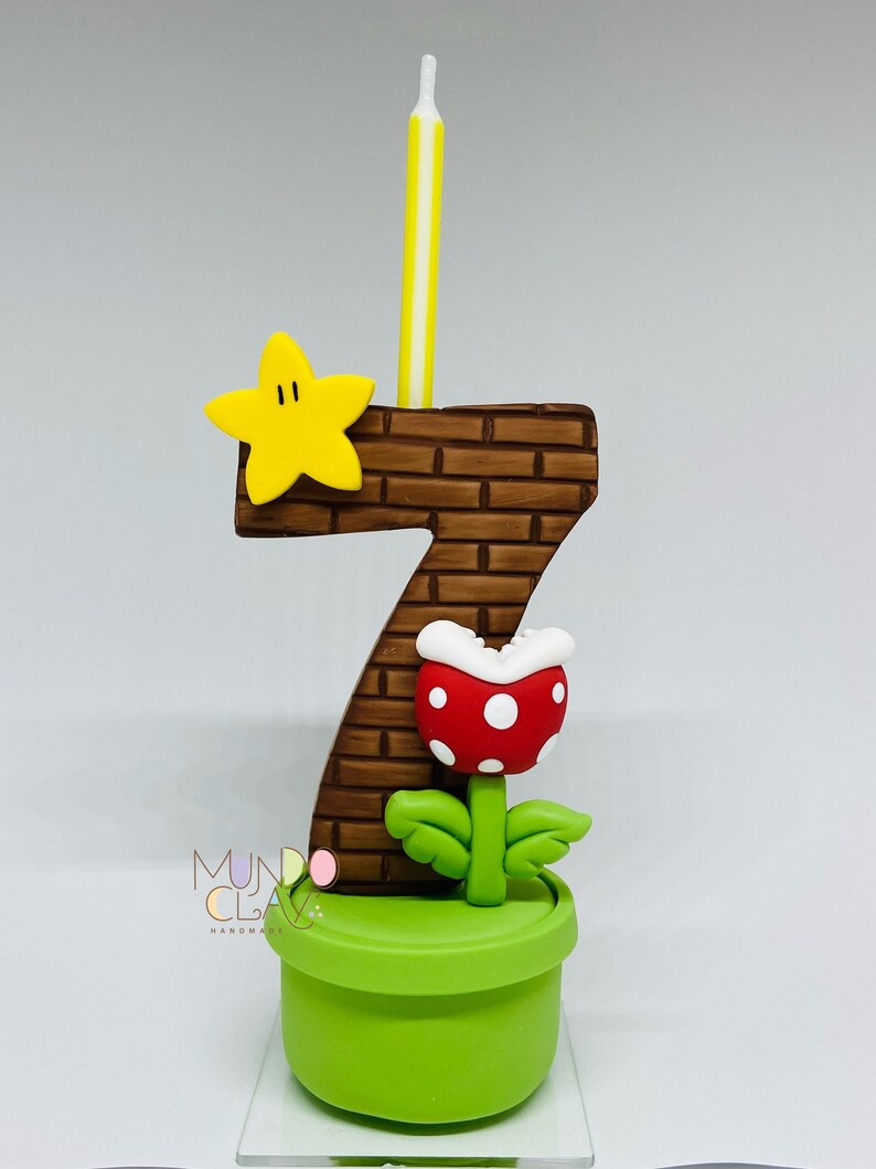 May include: A number seven birthday candle with a brick pattern, a yellow star, and a red and white Piranha Plant. The candle is yellow and white. The base is a green pot. The design is inspired by the Super Mario video game series.