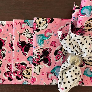 Puede incluir: Servilletas rosas con el tema de Minnie Mouse con un servilletero a juego. Las servilletas presentan un patr&oacute;n repetido de Minnie Mouse, corazones y lunares. El servilletero es plateado.