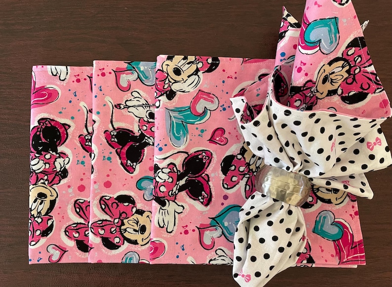 Puede incluir: Servilletas rosas con el tema de Minnie Mouse con un servilletero a juego. Las servilletas presentan un patr&oacute;n repetido de Minnie Mouse, corazones y lunares. El servilletero es plateado.