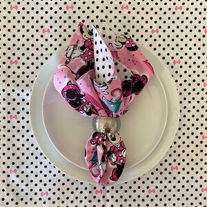 Puede incluir: Una servilleta rosa doblada con un estampado de Minnie Mouse, sujeta por un servilletero plateado, descansa sobre un plato blanco. La servilleta presenta un patr&oacute;n de lunares blancos y negros y un dise&ntilde;o de Minnie Mouse. El fondo es un mantel blanco con lunares negros y lazos rosas.