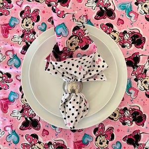 Puede incluir: Una servilleta doblada con un estampado de Minnie Mouse, sujeta por un servilletero plateado, descansa sobre un plato blanco. La servilleta presenta un fondo rosa con ilustraciones de Minnie Mouse y un estampado de lunares blancos y negros.
