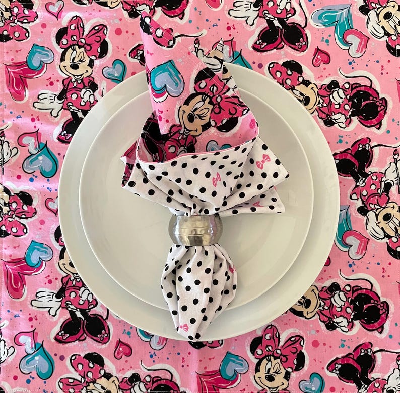 Puede incluir: Una servilleta doblada con un estampado de Minnie Mouse, sujeta por un servilletero plateado, descansa sobre un plato blanco. La servilleta presenta un fondo rosa con ilustraciones de Minnie Mouse y un estampado de lunares blancos y negros.
