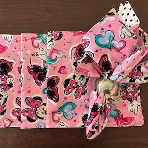 Puede incluir: Servilletas de tela rosas con un estampado de Minnie Mouse, con corazones y lunares. Una servilleta est&aacute; doblada y sujeta por un servilletero plateado. Las servilletas est&aacute;n sobre una superficie de madera marr&oacute;n.