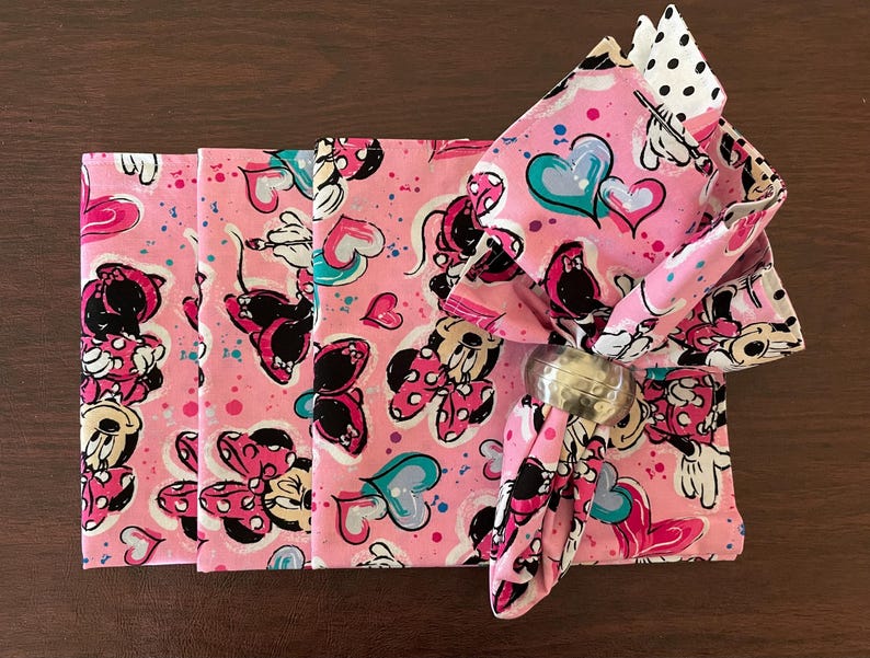 Puede incluir: Servilletas de tela rosas con un estampado de Minnie Mouse, con corazones y lunares. Una servilleta est&aacute; doblada y sujeta por un servilletero plateado. Las servilletas est&aacute;n sobre una superficie de madera marr&oacute;n.