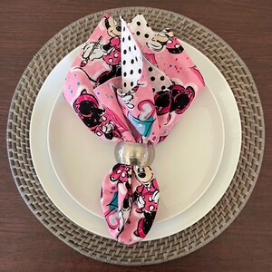 Puede incluir: Una servilleta rosa doblada con un estampado de Minnie Mouse, sujeta por un servilletero plateado, descansa sobre un plato blanco y un salvamanteles tejido. La servilleta tiene un estampado de lunares y un dise&ntilde;o de Minnie Mouse.