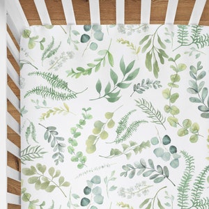 May include: A white fitted crib sheet with a green watercolor leaf pattern. The pattern features various types of leaves, including ferns, eucalyptus, and other foliage.