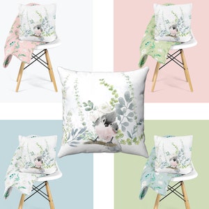 May include: A set of four throw pillows with a watercolor design of two birds perched on a branch with green leaves. The pillows are displayed on white chairs with floral patterned throw blankets.