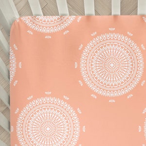 May include: A peach-colored fitted crib sheet with a white mandala pattern. The sheet is made of soft, breathable fabric and is perfect for a nursery.