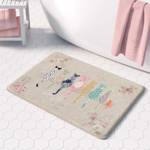 May include: A rectangular bath mat with a beige background featuring a row of seven stylized cats in various colors and patterns, including black, pink, orange, and teal. The mat is decorated with faded floral motifs and script text.