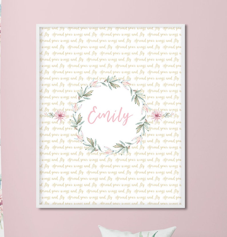 May include: A framed print with a light pink background and gold text repeating the phrase "spread your wings and fly." The center features a floral wreath with the name "Emily" in pink script.