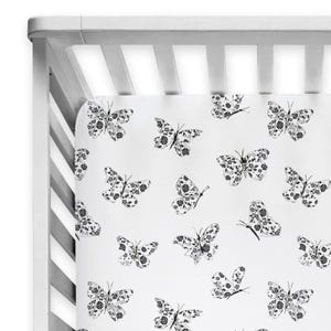 May include: A white crib sheet featuring a repeating pattern of black butterflies. Each butterfly is decorated with intricate floral details on its wings. The sheet is fitted over a white wooden crib frame with vertical slats visible at the top.