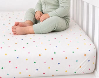 May include: A white crib sheet featuring a colorful polka dot pattern in shades of red, blue, green, yellow, and pink. The sheet is fitted over a mattress inside a white crib, with a child wearing a sage green long-sleeved onesie sitting on top.