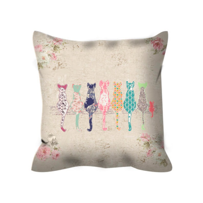 May include: A decorative pillow with a beige background and a row of colorful cats sitting on a branch. The cats are all different colors and patterns, and the pillow has a floral design around the edges.