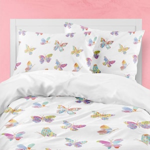 Könnte beinhalten: Ein weißer Bettbezug und zwei passende Kissenbezüge mit einem sich wiederholenden Muster aus bunten Schmetterlingen. Jeder Schmetterling zeigt ein Aquarell-Design mit verschiedenen Farben wie Blau, Gelb, Rosa und Violett auf dem Stoff.
