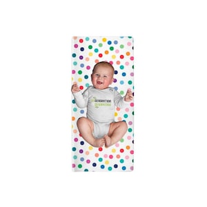 May include: A white baby bodysuit with long sleeves featuring the text REGENERATION GENERATION in black and green lettering. The garment is displayed against a white background covered in a repeating pattern of multicolored polka dots.