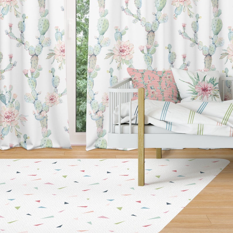 May include: A bedroom scene featuring a white crib, curtains, and a rug. The curtains and a pillow display a repeating pattern of watercolor cacti and pink flowers. The rug has a white background with colorful triangle shapes.