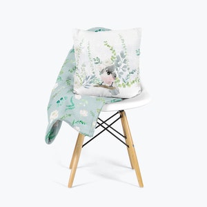 May include: A light blue throw blanket with a floral pattern featuring white flowers and green leaves. A white chair with wooden legs has a throw pillow with a watercolor illustration of a bird perched on a branch with green leaves.